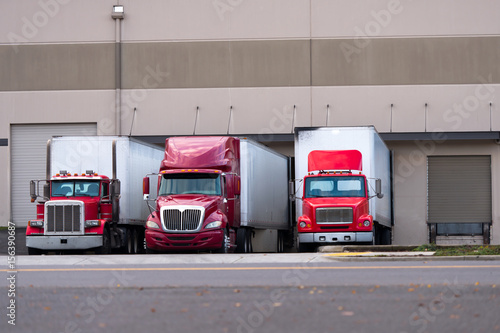 Slika na platnu Three red semi trucks are among the dock for loading trailers