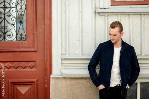 Portrait of a blonde man wearing a blue leather jacket, leaning back against a wall of a wooden building on a sunny day.