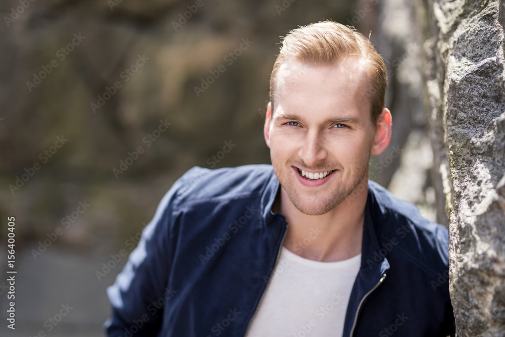An attractive man standing leaning against a wall of stone wearing a blue jacket and white shirt, feeling relaxed and confident on a sunny day. An attractive man standing leaning against a wall of stone wearing a blue jacket and white shirt, feeling relaxed and confident on a sunny day.