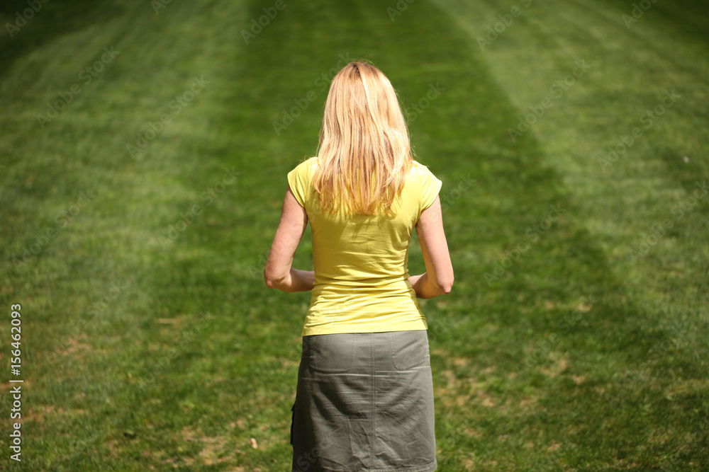 Thoughtful sad blonde long hair woman stand alone in the sunny park. View from back. Thoughtful sad blonde long hair woman stand alone in the sunny park. View from back.