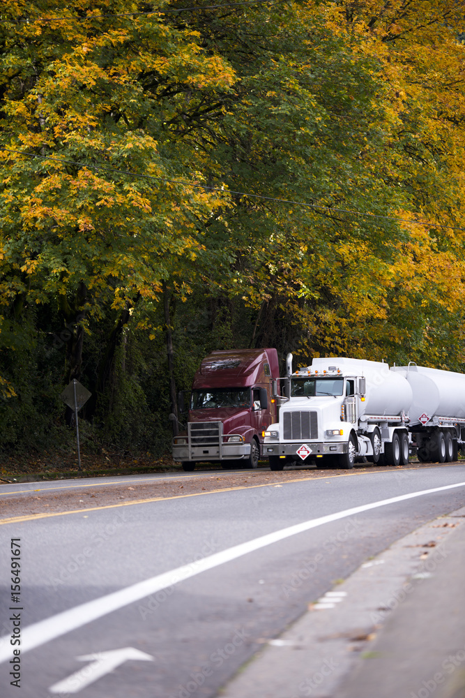 Two big rigs semi trucks with trailers on autumn road with yellow trees