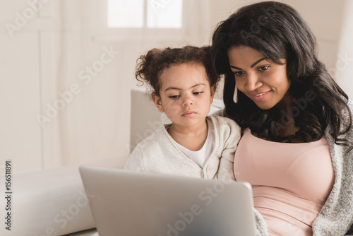 Beautiful mother and daughter sitting on couch and using laptop together