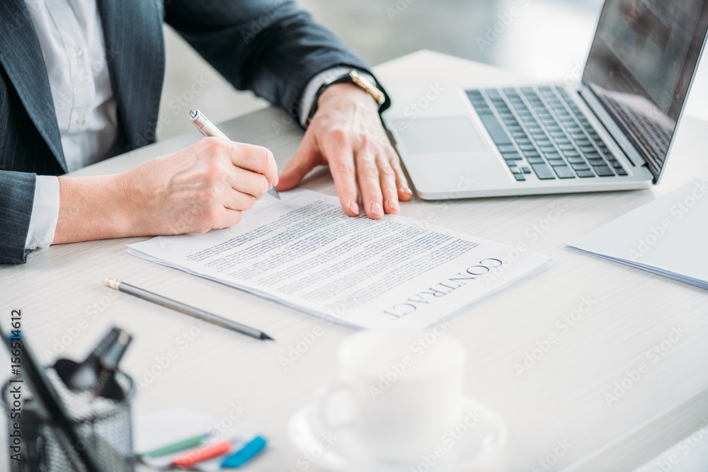 partial view of businesswoman sitting at workplace and signing contract partial view of businesswoman sitting at workplace and signing contract