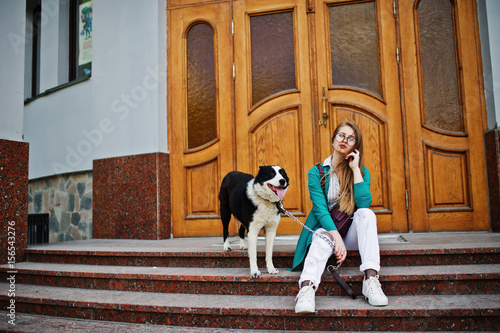 Trendy girl at glasses and ripped jeans with russo-european laika (husky) dog on a leash, against wooden door on street of city. Friend human with animal theme.
