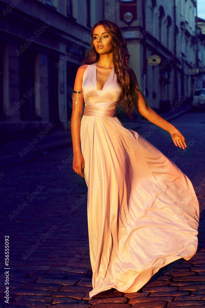 fashion portrait of young beautiful girl in pale lilac color long flying dress walking down the street in old town at sunset      