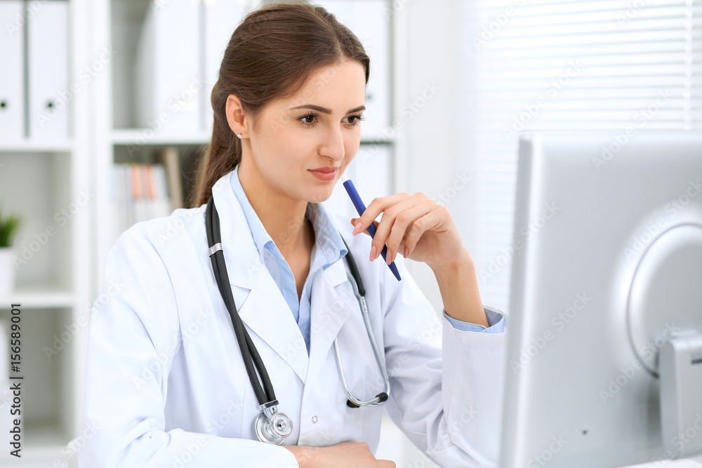Latin american female doctor sitting at the table and working by computer at hospital office. The physician or therapist makes a diagnosis. Health care, medicine and patient service concept Latin american female doctor sitting at the table and working by computer at hospital office. The physician or therapist makes a diagnosis. Health care, medicine and patient service concept