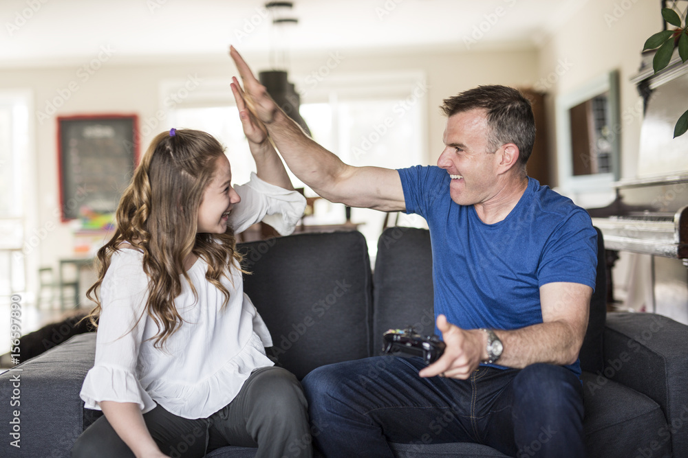 Fototapeta premium Handsome father and his cute little daughter are playing game console and smiling while sitting on couch at home.