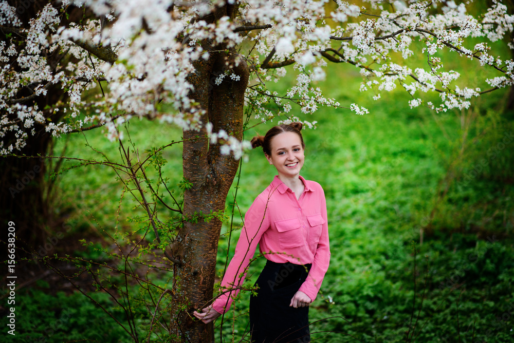 Young beautiful girl in a pink shirt standing under blossoming apple tree and enjoying a sunny day.