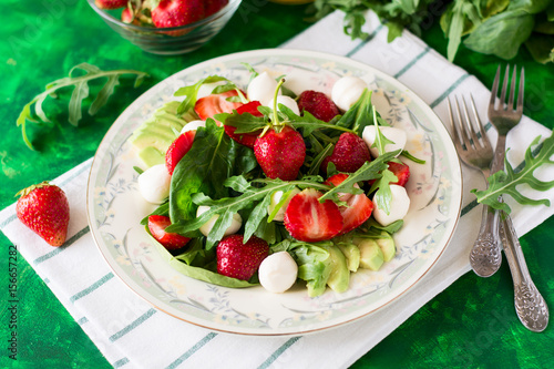 Fresh vegetarian salad with spinach, arugula, avocado slices, strawberries and mini mozzarella on green wooden table. Selective focus