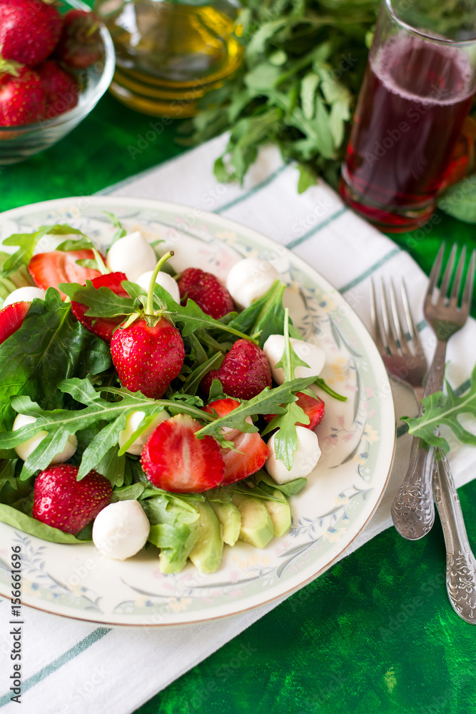 Fresh vegetarian salad with spinach, arugula, avocado slices, strawberries and mini mozzarella on green wooden table. Selective focus