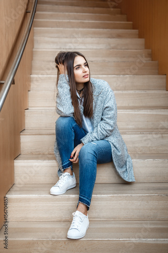 Portrait Of Pensive Caucasian Brunette Young Beautiful Girl Woman With Long Dark Hair And Brown Eyes In White Shirt Grey Coat And Blue Jeans Sitting On Staircase Looking Away Stock Photo Portrait Of Pensive Caucasian Brunette Young Beautiful Girl Woman With Long Dark Hair And Brown Eyes In White Shirt Grey Coat And Blue Jeans Sitting On Staircase Looking Away Stock Photo