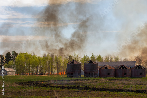 A wall of fire with billowing smoke coming toward a line of green leaf trees and old abandoned wood grain bins in a agricultural summer landscape