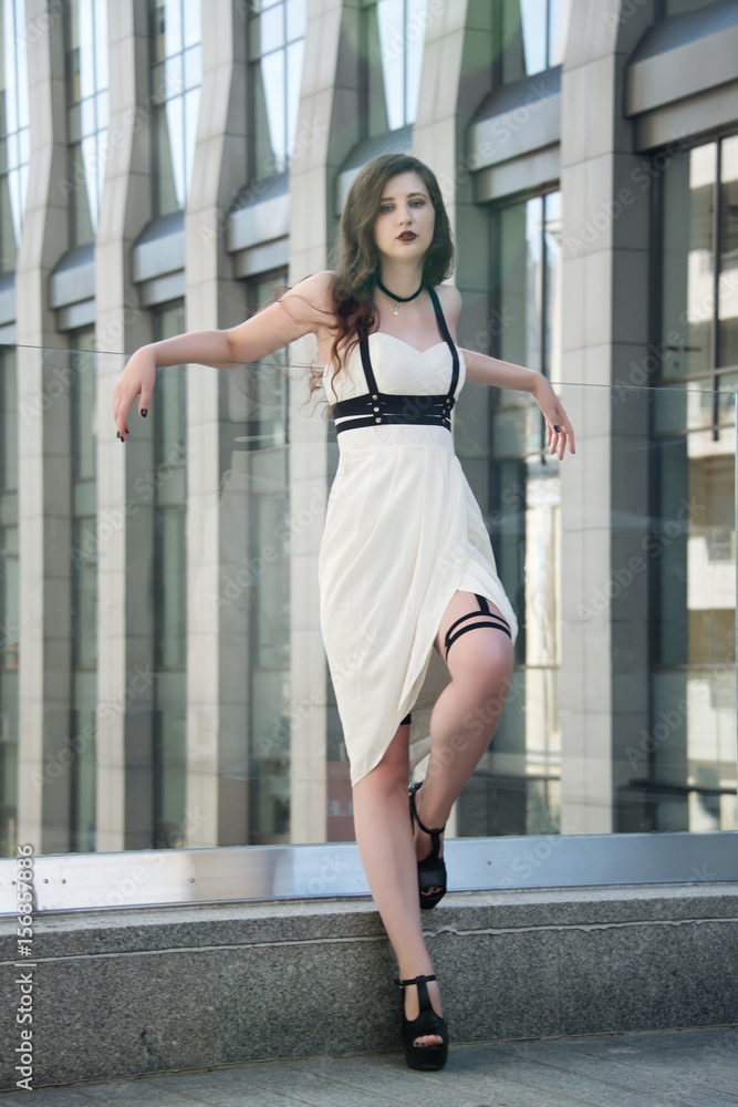 Young beautiful sexy woman wearing trendy outfit, white dress and leather swordbelt. Longhaired brunette posing in the city street. Outdoor fashion photography Young beautiful sexy woman wearing trendy outfit, white dress and leather swordbelt. Longhaired brunette posing in the city street. Outdoor fashion photography