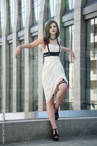 Young beautiful sexy woman wearing trendy outfit, white dress and leather swordbelt. Longhaired brunette posing in the city street. Outdoor fashion photography
