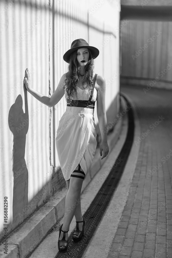 Obraz premium Young beautiful sexy woman wearing trendy outfit, white dress, black hat and leather swordbelt. Longhaired brunette posing in the city street. Outdoor fashion photography