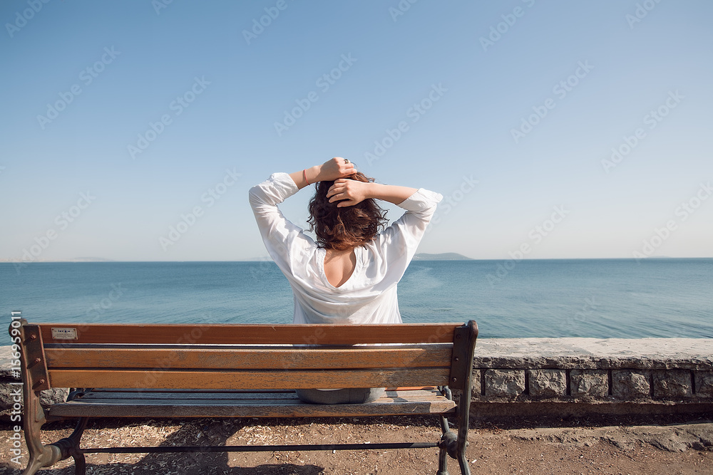 Back portrait woman sitting on a bench at the sea.amazing view,girl with curly hair,woman in white shirt,resting,spring holidays,summer time,relax,take it easy,trendy,summer concept,girl relaxing