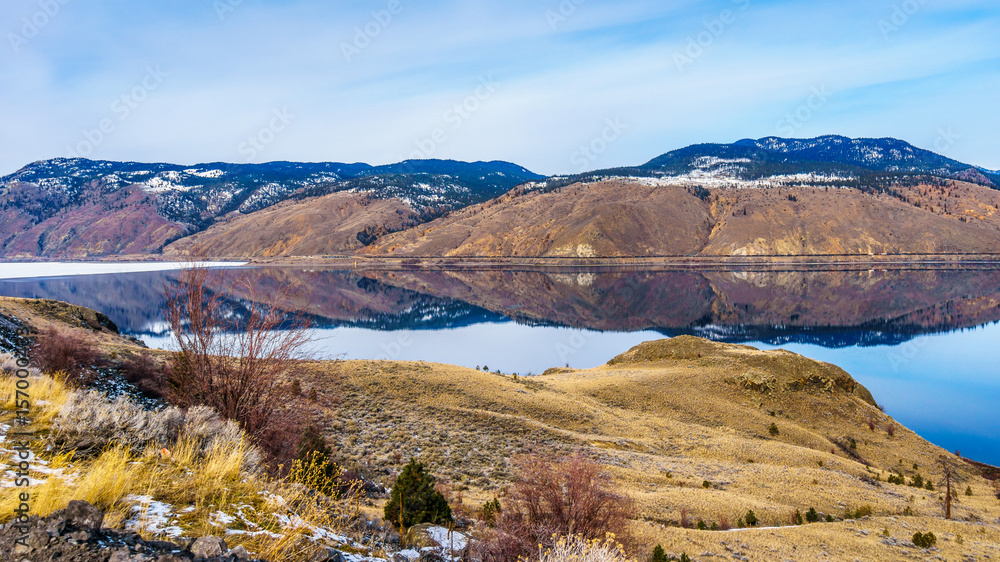 Kamloops Lake, which is a very wide portion of the Thompson River, on a cold winter day with the surrounding mountain reflecting in the quiet surface of the lake. 