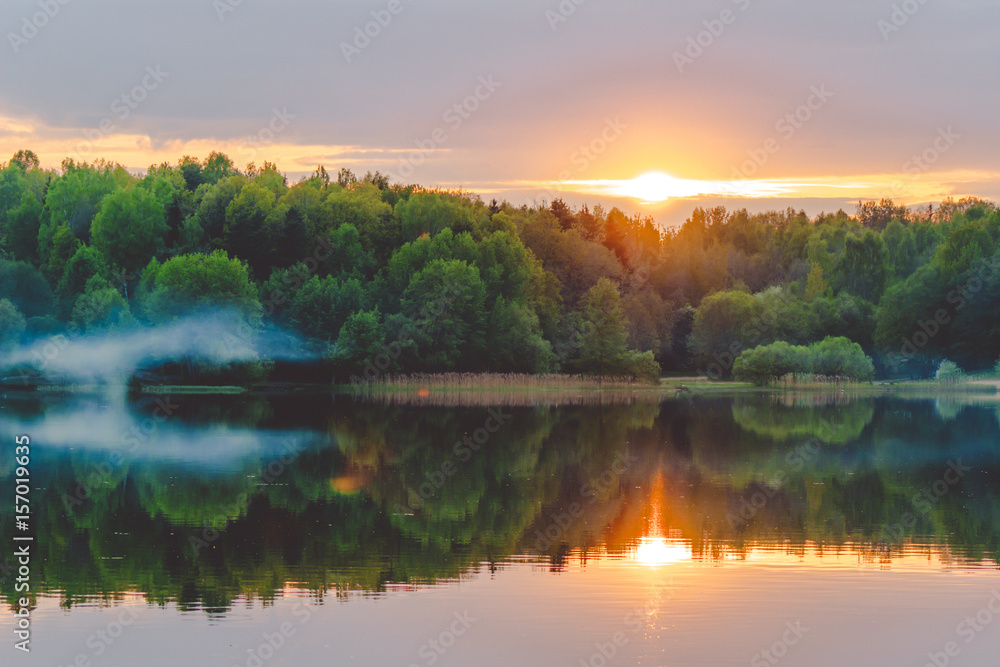 A calm sunset on the shore of the lake, where the forest reflects in calm water with haze and fog