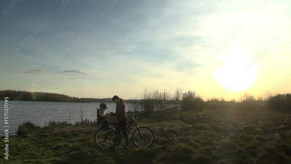 A young woman in a spotted sweatshirt puts a child in a striped sweater and a protective helmet in a bike child seat on the street next to the pond on a summer evening at sunset. A young woman in a spotted sweatshirt puts a child in a striped sweater and a protective helmet in a bike child seat on the street next to the pond on a summer evening at sunset.