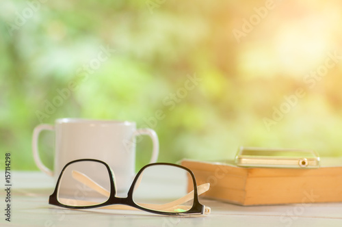 White cup coffee with glasses and book on white table against nature background