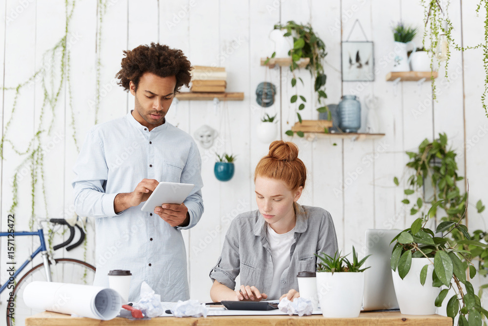 Team work of hard-working architect workers. Attractive male with trendy hairdo holding tablet and pretty redhead woman dressed in casual clothes making calculations isolated over home cozy interior