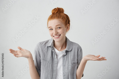 Studio portrait of attractive redhead freckled teenager having broad smile shrugging her shoulders having some doubts. Emotional woman with red bun being confused while making some decisions