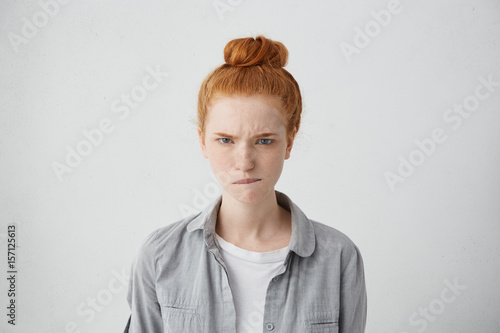 Portrait of angry frowning teenager having ginger bun biting her lip with anger isolated over white background. Young freckled redhead woman having dissatisfied expression after hearing bad news