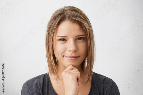 Close-up portrait of cute young female with trendy hairdo and pure skin wearing casual clothes holding hand on cheek looking suspicious into camera posing against white background. People, lifestyle