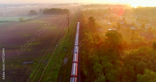 Freight train rolls through foggy rural landscape at sunrise, aerial view, breathtaking scenic beauty.
