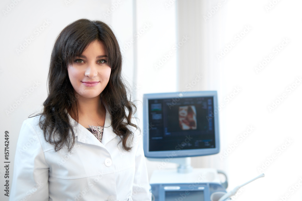 Portrait of professional female nurse in white uniform at modern and new clinic, standing at cabinet, posing, smiling at camera. Attractive brunette doctor against ultrasound equipment and computer. Portrait of professional female nurse in white uniform at modern and new clinic, standing at cabinet, posing, smiling at camera. Attractive brunette doctor against ultrasound equipment and computer.
