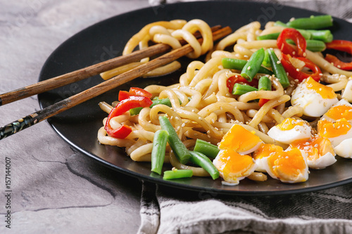 Cooking stir fry udon noodles, green beans, sliced paprika, boiled eggs, soy sauce with sesame seeds in black plate with wood chopsticks over gray texture background. Asian style dinner close up