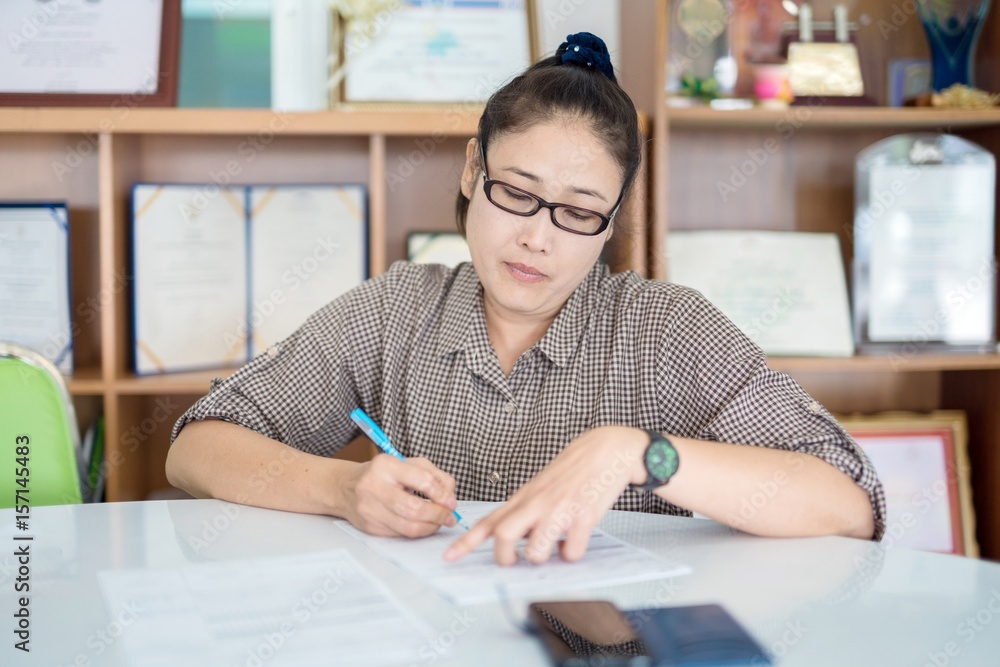 middle aged asian woman wear glasses working at her desk taking notes at office. middle aged asian woman wear glasses working at her desk taking notes at office.