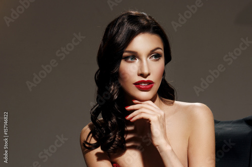 Young pretty beautiful woman in red long evening dress with makeup  red lips and classical hollywood waves hairstyle sitting in director s chair at film set with two cinema lights.