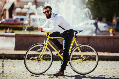 Handsome hipster young bearded man looking at camera while sitting on his bicycle outdoors near fountains