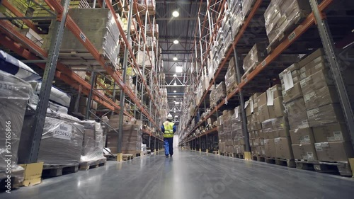 Back view. Female warehouse worker walks through rows of storage racks with merchandise