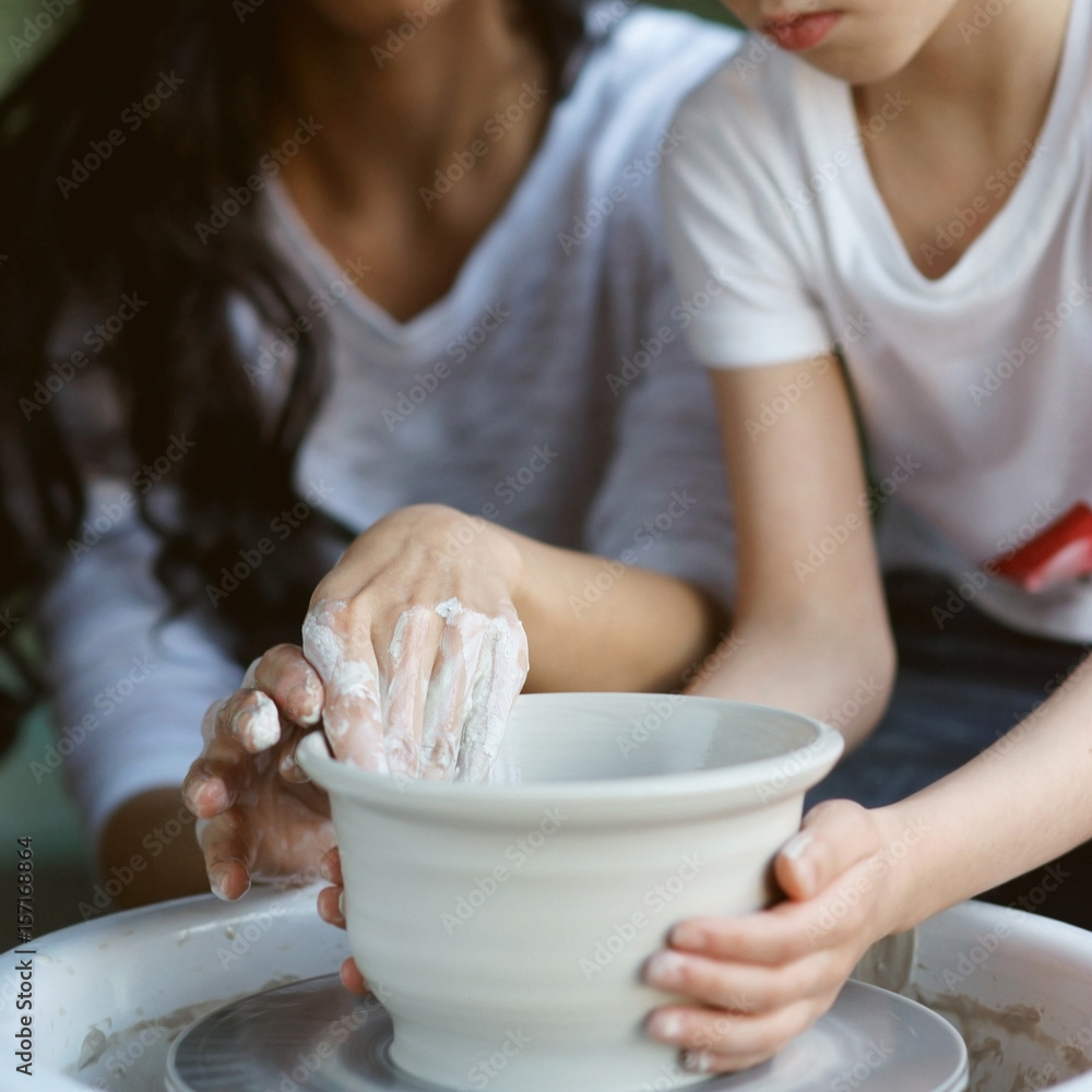 Pottery classes. Young beautiful pretty caucasian brunette mother teaching her son. Son making clay pot on potter wheel. Shallow DOF. Focus on woman hands.
