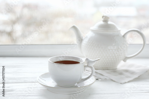 Cup of hot drink with teapot on wooden windowsill