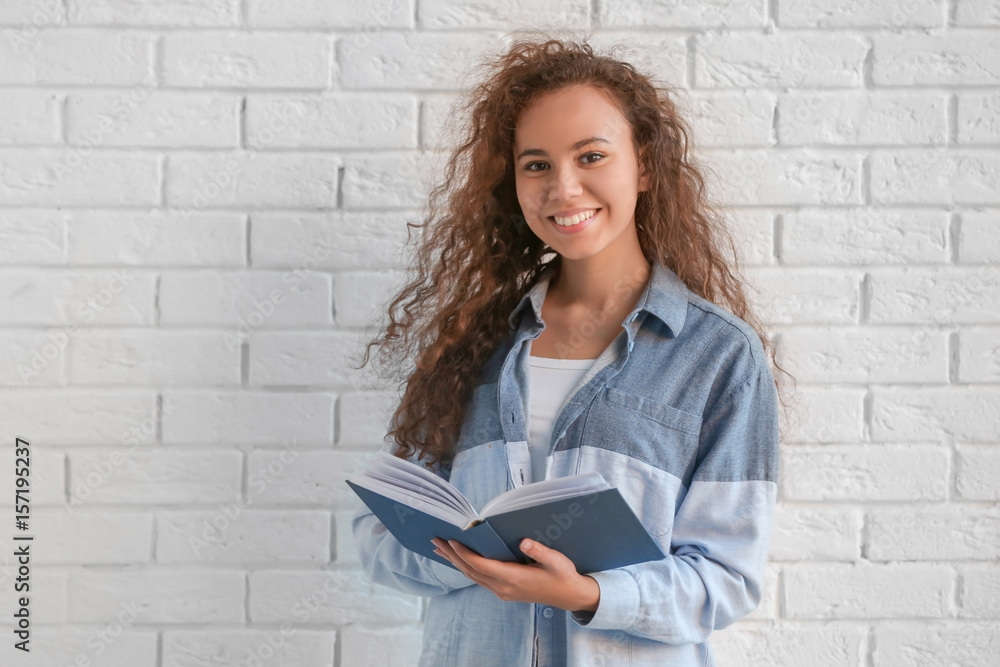 Beautiful young African American woman with book standing near brick wall Beautiful young African American woman with book standing near brick wall