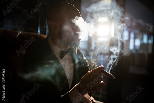 A brutal man in a dress coat smokes a cigar.