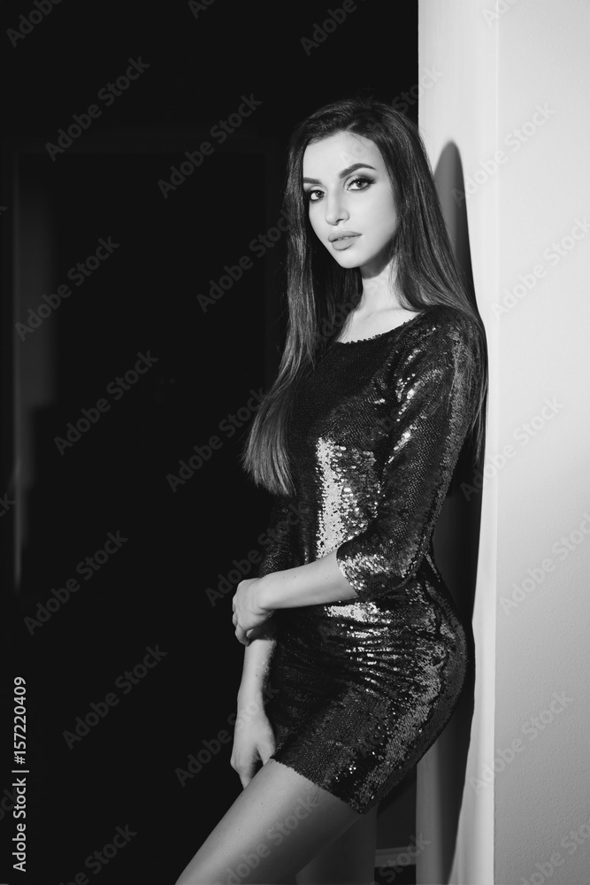 Fototapeta premium Young sexy girl wearing blue green shiny dress with sequins standing in inreior near wall. Fashion model with long brunette hair and makeup