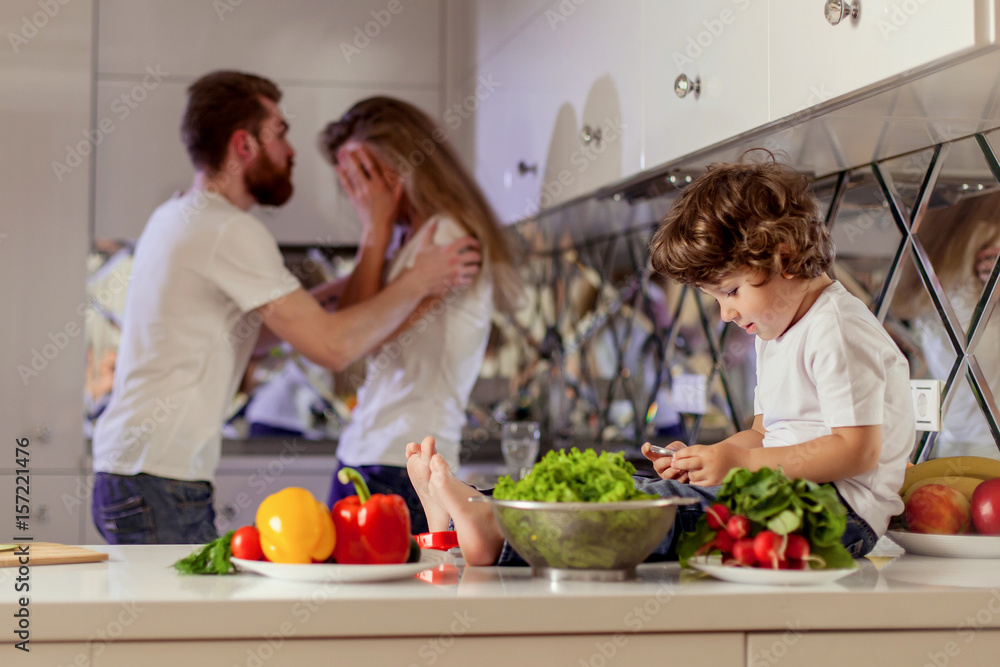 Small boy with the phone in his hands sitting on the kitchen surface while his parents fighting on the background. Small boy with the phone in his hands sitting on the kitchen surface while his parents fighting on the background.