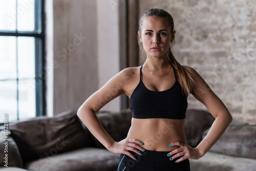 Portrait of strong slim sporty Caucasian young sportswoman wearing black sports bra standing with hands on hips looking at camera in loft studio