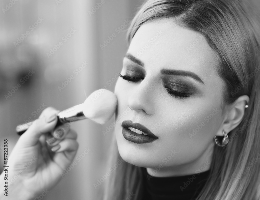 Obraz premium Process of making makeup. Mak-up artist working with brush on model face. Portrait of young blonde woman in beauty saloon interior. Applying tone to skin.