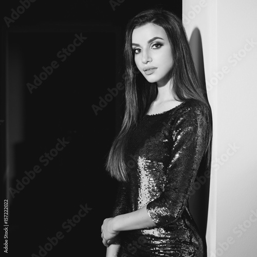 Young sexy girl wearing blue green shiny dress with sequins standing in inreior near wall. Fashion model with long brunette hair and makeup photo
