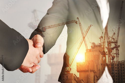 Double exposure of handshake, construction crane, building and sunset in the evening as business, relationship, partnership, commitment and industry concept.