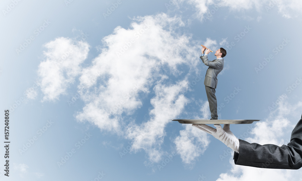 Businessman on metal tray playing fife against blue sky background Businessman on metal tray playing fife against blue sky background