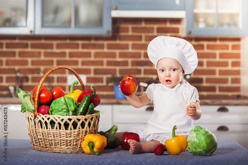 Little girl dressed in white chef hat and apron sits among vegetables and holds an apple. Brick kitchen wall at the background. One year old child nutrition. Horizontal Little girl dressed in white chef hat and apron sits among vegetables and holds an apple. Brick kitchen wall at the background. One year old child nutrition. Horizontal