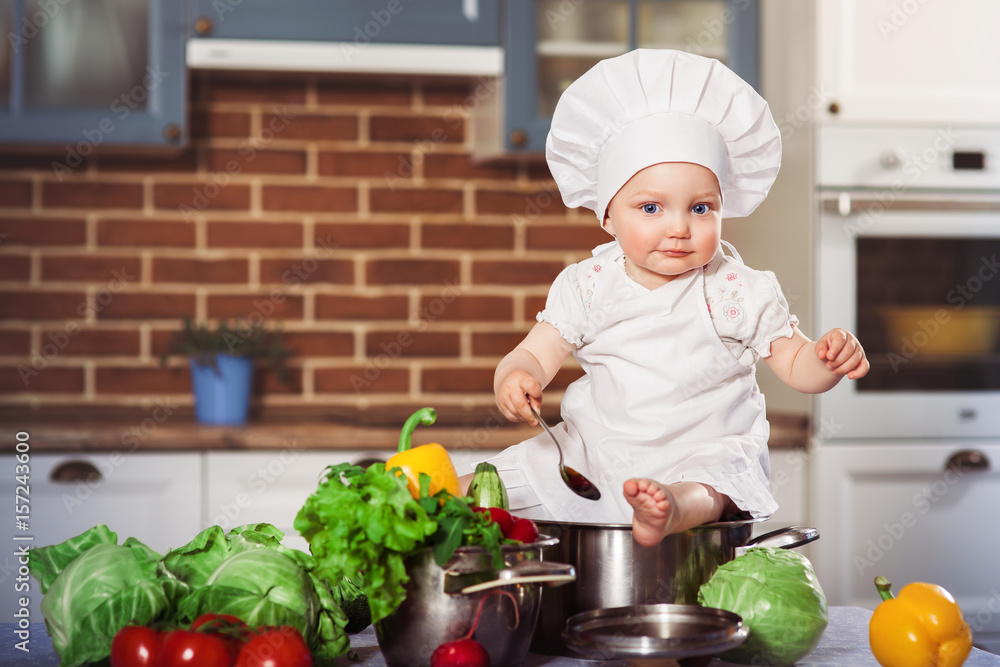 Baby girl dressed in white chef toque and apron baby sitting with spoon astride a stainless pan filled by raw vegetables. Brick wall background. One year old child nutrition. Horizontal Baby girl dressed in white chef toque and apron baby sitting with spoon astride a stainless pan filled by raw vegetables. Brick wall background. One year old child nutrition. Horizontal