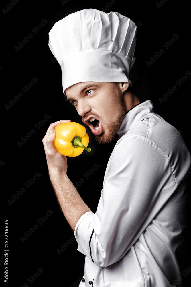 Young bearded man chef In white uniform holds yellow bell pepper on black background Young bearded man chef In white uniform holds yellow bell pepper on black background