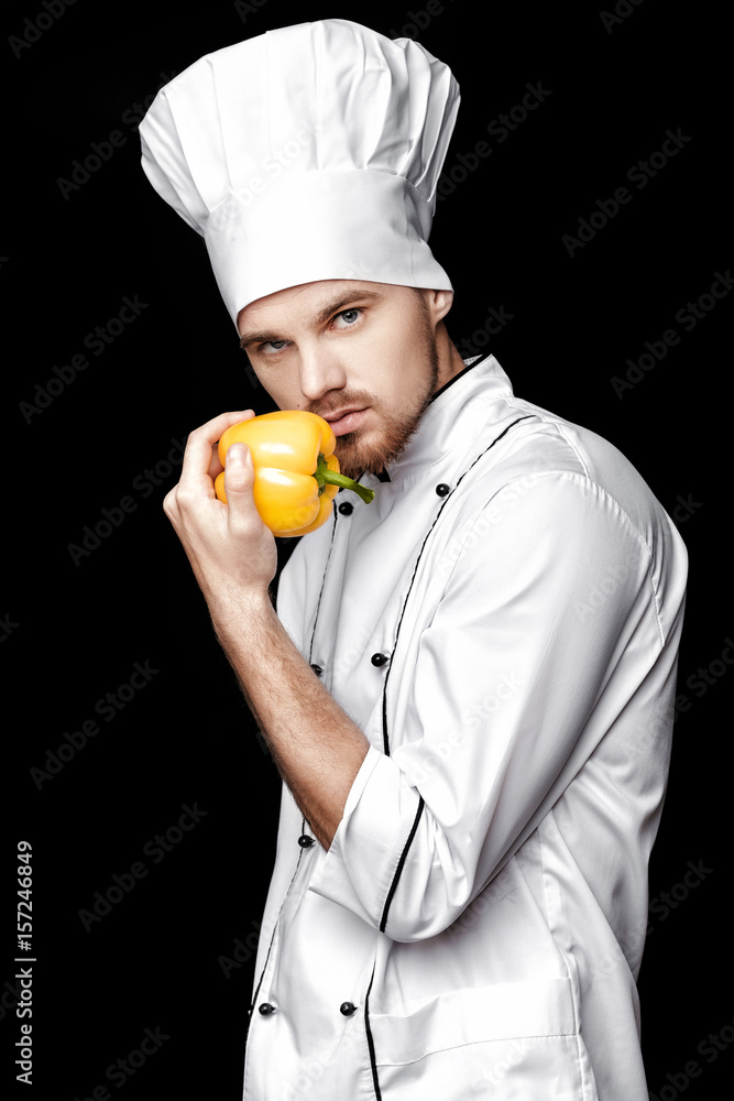 Young bearded man chef In white uniform holds yellow bell pepper on black background Young bearded man chef In white uniform holds yellow bell pepper on black background